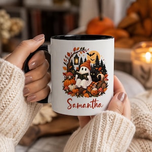 May include: A white ceramic mug with a black handle and rim, featuring a colorful autumn-themed illustration of a ghost reading a book, a black cat, pumpkins, mushrooms, and a lantern. The name Samantha is printed in orange cursive text below.