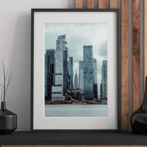 Può includere: Stampa incorniciata con un paesaggio urbano moderno con grattacieli. L'immagine mostra una vista sul lungomare con un cielo nuvoloso. La stampa è in una cornice nera, aggiungendo un tocco di eleganza alla scena.