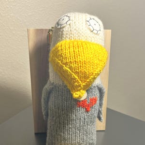 May include: Hand-knitted bird doll with a white head, yellow beak and legs, and a gray body. The doll has embroidered eyes and a red heart-shaped design on the chest. The bird is standing on a gray surface.
