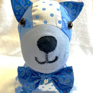 May include: A handmade stuffed dog with a blue and white patchwork design. The dog has a black nose, button eyes, and a blue bow tie. The ears and bow tie feature a paisley pattern.