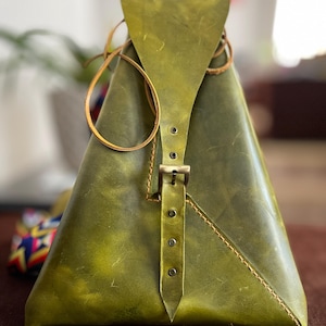 May include: An olive green leather bag in a triangular shape, with a flap and buckle closure. The bag has a stitched design and a leather strap. The bag's design is unique.