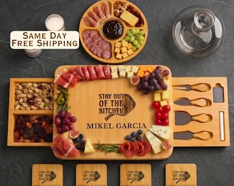 May include: A rectangular bamboo charcuterie board with slide-out drawers containing cheese knives, featuring the engraved text "STAY OUT OF THE KITCHEN MIKEL GARCIA". Includes a round serving tray, four square coasters, and assorted food items.