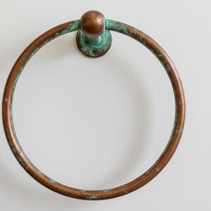 May include: A round, antique-style towel ring made of copper. The ring has a weathered appearance with areas of green patina. It is mounted on a circular base and is designed for wall mounting.