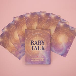 Puede incluir: Un mazo de cartas or&aacute;culo dispuesto en abanico detr&aacute;s de una caja central. Las cartas y la caja presentan un dise&ntilde;o c&oacute;smico en violeta y dorado con patrones de luz circulares. La caja muestra el texto BABY TALK y Messages from the Unborn Child de Dionne Douball.
