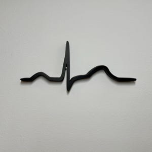 Könnte beinhalten: Ein schwarzes Metall-Wandkunstwerk in Form eines Elektrokardiogramms (EKG) oder einer Herzschlaglinie. Das Kunstwerk ist an einer weißen Wand befestigt und schafft eine minimalistische und moderne Ästhetik. Das Design ist eine einfache, aber eindrucksvolle Darstellung eines Herzschlags.
