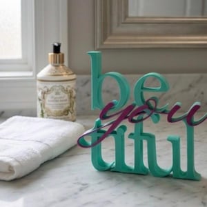 Könnte beinhalten: Ein türkisfarbenes und magentafarbenes dekoratives Schild "be you tiful" steht auf einer Marmorarbeitsplatte. Ein weißes Handtuch und eine Flasche Lotion befinden sich ebenfalls auf der Theke. Das Schild ist ein dekorativer Artikel für ein Badezimmer oder Schlafzimmer.