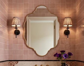 May include: Interior of a bathroom with a wooden vanity unit, marble countertop, and decorative mirror. The walls are tiled in pink, and the floor has a patterned design. Sconces flank the mirror. A vase of purple flowers adds a touch of colour.