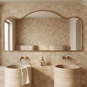 Pode incluir: Uma casa de banho moderna com uma parede de azulejos bege e um grande espelho com moldura dourada. Duas pias cilíndricas com torneiras douradas estão posicionadas sob o espelho. Um dispensador de sabão está posicionado entre as pias. Uma toalha branca está pendurada numa pia.
