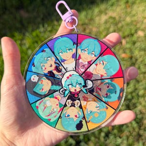 May include: A clear, circular keychain featuring a colorful anime-style design. The design is divided into segments, each showcasing a different character with blue hair and various outfits. A purple clasp is attached at the top.