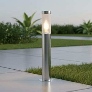 May include: A contemporary outdoor lamp featuring a frosted glass cylinder and a silver metal base. The lamp is lit, providing a warm light. It stands on a concrete patio, with grass and plants in the background.