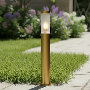 May include: A gold-coloured outdoor pathway light with a cylindrical design. The light has a frosted glass top and a warm, glowing bulb. The light is set on a stone pathway with green grass and a garden in the background.