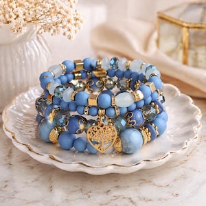 Peut inclure: Plusieurs bracelets de perles bleues avec des accents dorés et un charm en forme de cœur doré. Les bracelets présentent différentes nuances de perles bleues, dont des perles mates et en cristal. Les bracelets sont présentés sur une assiette blanche à bords festonnés.