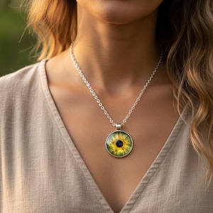 Peut inclure: Un collier en argent avec un pendentif rond représentant un tournesol. Le pendentif présente un motif floral jaune et vert avec un centre sombre. Le collier est porté sur un haut de couleur neutre.