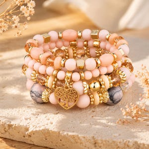 Può includere: Set di cinque braccialetti impilati in tonalità rosa, pesca e oro. I braccialetti presentano una varietà di perline, tra cui forme rotonde, ovali e quadrate, con un ciondolo a forma di cuore dorato. I braccialetti sono impilati insieme.