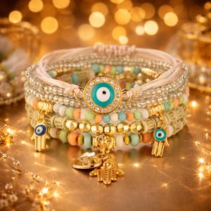 Peut inclure: Plusieurs bracelets de perles aux couleurs variées, dont blanc, or, turquoise et pêche. Les bracelets comportent des breloques avec un motif d'œil protecteur et une main de Hamsa dorée. Les bracelets sont sur une surface dorée.