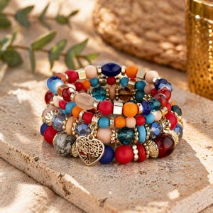 Peut inclure: Plusieurs bracelets de perles multicolores, rouges, bleues, oranges et crème. Les bracelets présentent un mélange de perles rondes et facettées, avec un charm en forme de cœur doré. Les bracelets sont empilés sur une surface en pierre.