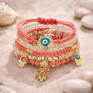 Peut inclure: Plusieurs bracelets de perles corail et dorées avec des breloques. Les bracelets présentent un charme d'œil maléfique, des breloques de main de Hamsa et divers motifs de perles. Les bracelets sont exposés sur une surface sablonneuse avec des coquillages et des pierres.