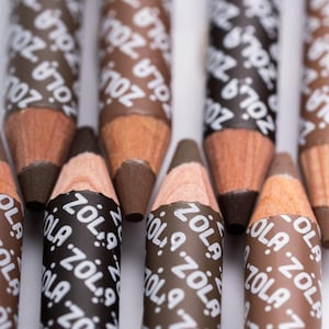 以下が含まれることがあります： 茶色と黒の眉ペンシルのクローズアップ。鉛筆は木製のボディで、「ZOLA」と白で書かれたラベルが巻かれています。鉛筆の先端はさまざまな茶色の色合いです。