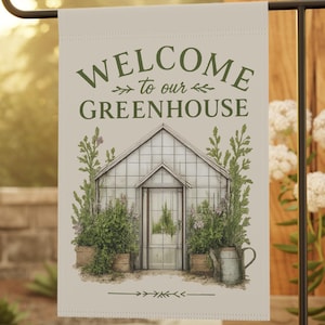 以下が含まれることがあります： 緑色の文字で「WELCOME to our GREENHOUSE」と書かれたガーデンフラッグ。温室、植物、じょうろの詳細なイラストが特徴です。旗は庭に設置された黒い金属製のスタンドに表示されています。