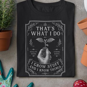 May include: A black t-shirt featuring a white graphic print of a hand holding a small plant. The text reads: THAT'S WHAT I DO, I GROW STUFF AND I KNOW THINGS, QUALITY SEEDS, STRONG ROOTS. The design is enclosed in a decorative rectangular border.