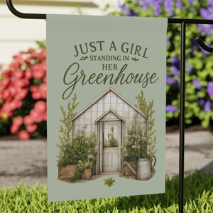 以下が含まれることがあります： 「JUST A GIRL STANDING IN HER Greenhouse」のテキストがスクリプトフォントで書かれたガーデンフラッグ。温室と植物、じょうろの水彩イラストが特徴です。黒い金属製のスタンドに吊るされています。