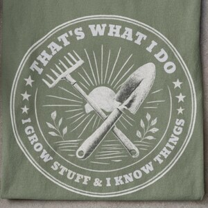 May include: Sage green t-shirt with a circular graphic featuring gardening tools and the text "THAT'S WHAT I DO I GROW STUFF & I KNOW THINGS." Gardening gloves, a potted plant, and red polka dot rain boots are also visible.