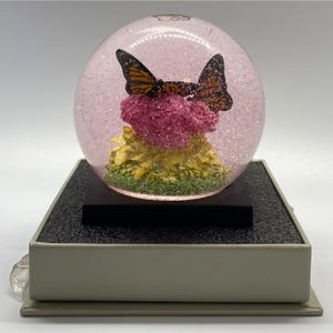 May include: A decorative snow globe featuring two orange and black butterflies perched atop a pink and yellow floral arrangement. The globe is filled with glitter and sits on a black base, which is set on a gray rectangular box.