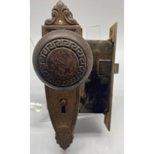 May include: Antique bronze door hardware set. The set includes a round doorknob with a Greek key design, a keyhole plate, and a rectangular latch mechanism. The hardware has a weathered appearance, suggesting age and use. The overall design is ornate and decorative.