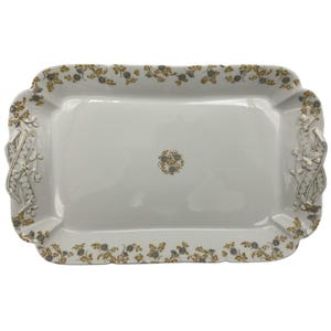May include: A rectangular white ceramic serving platter with a floral design in gold and gray. The edges are scalloped, and the handles are decorated with a silver floral relief. A small floral design is in the center.