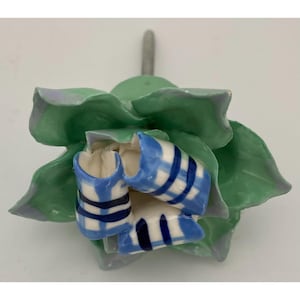 May include: A ceramic floral sculpture with green petals edged in lavender and blue and white striped accents. The sculpture has a silver-colored stem. The overall design is reminiscent of a flower.