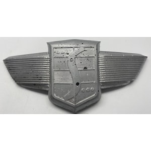 May include: A silver-colored emblem featuring a central shield design with a curved vertical line, horizontal lines, and small circular and triangular accents. The shield is flanked by horizontal wings on both sides, all with a speckled finish.
