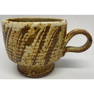 May include: A ceramic mug with a speckled cream and brown finish, featuring a textured surface with vertical brown stripes. The mug has a rounded bowl shape, a pedestal base, and a curved handle, all displaying a rustic, hand-crafted appearance.