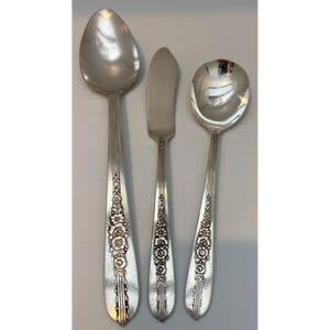 May include: A set of three silver-toned metal utensils consisting of a large oval spoon, a flat-edged butter knife, and a smaller round soup spoon. Each handle features a vertical ribbed design and an embossed floral pattern.