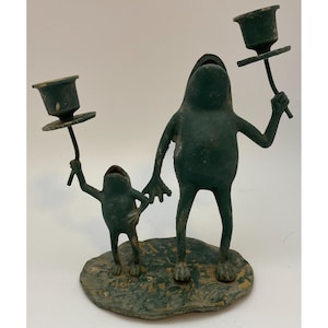 May include: A whimsical, dark green frog-themed candle holder. Two frog figures, one large and one small, stand on a lily pad base, each holding a candle cup. The metal sculpture has a distressed finish.