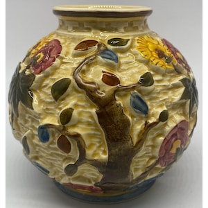 May include: A round ceramic vase with a cream-colored background featuring a raised, embossed design of a brown tree trunk, multi-colored leaves in shades of green, blue, and brown, and large red and yellow flowers with a textured surface.
