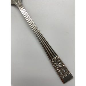 May include: A silver spoon with a decorative floral design on the handle. The spoon's handle has vertical grooves and a rectangular end with an embossed floral pattern. The spoon is made of a shiny, reflective metal.