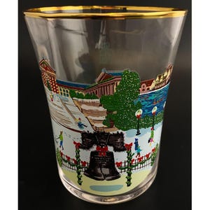 May include: Clear glass with a gold rim. The glass features a colourful winter scene with the Liberty Bell and a snowy landscape. The design includes buildings, trees, and people, creating a festive holiday theme.