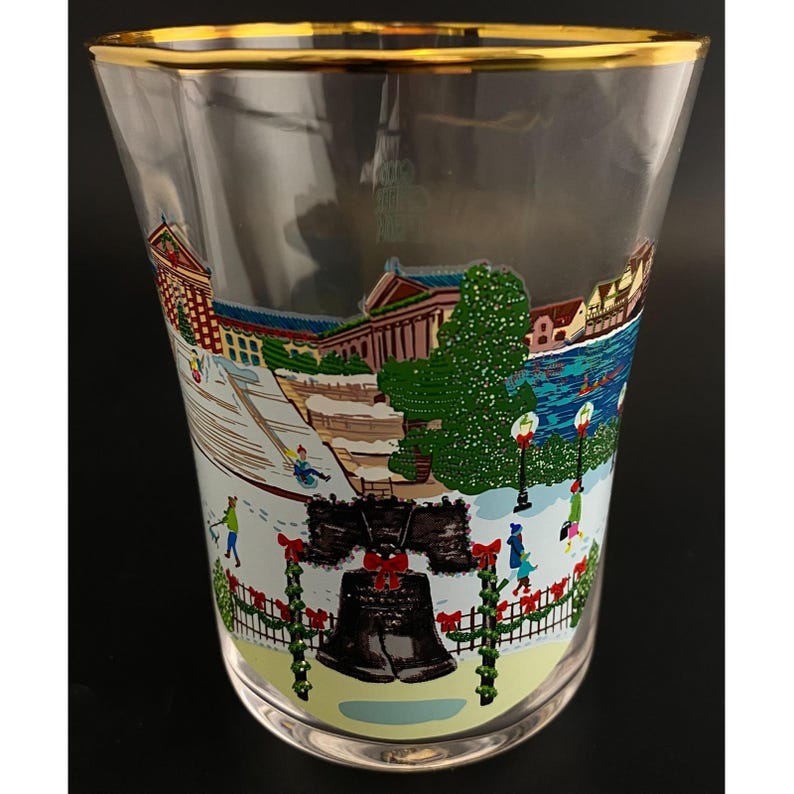 May include: Clear glass with a gold rim. The glass features a colourful winter scene with the Liberty Bell and a snowy landscape. The design includes buildings, trees, and people, creating a festive holiday theme.