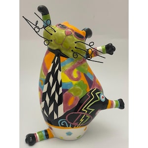 May include: A ceramic cat figurine featuring a vibrant, multi-colored abstract pattern of yellow, orange, blue, and pink. The cat wears a black and white chevron-patterned tie, has a lime green face, and includes black wire whiskers.