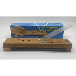 May include: A wooden knife sharpener with a clear plastic guide and a blue box in the background. The sharpener has a rectangular wooden base with a metal sharpening rod. The box reads "Ceramic Sharp".