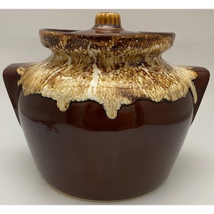 May include: A brown ceramic bean pot with a lid, featuring a wide, rounded body and two side handles. The upper portion of the pot and the lid are decorated with a textured, cream-colored drip glaze that contrasts against the smooth, dark brown base.