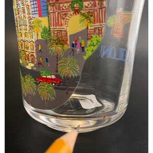 K&ouml;nnte beinhalten: Ein klares Glas mit einer farbenfrohen Illustration einer Stadtlandschaft, einschlie&szlig;lich Palmen, Geb&auml;uden und Menschen. Ein rotes Auto mit einem Weihnachtsbaum ist ebenfalls abgebildet. Das Glas ist zylindrisch und vermutlich ein Trinkglas.