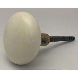 May include: A round, smooth, off-white porcelain door knob featuring a brass-toned metal collar with a visible screw. A dark, rectangular metal spindle extends from the back of the knob, showing three small circular holes at the end of the shaft.