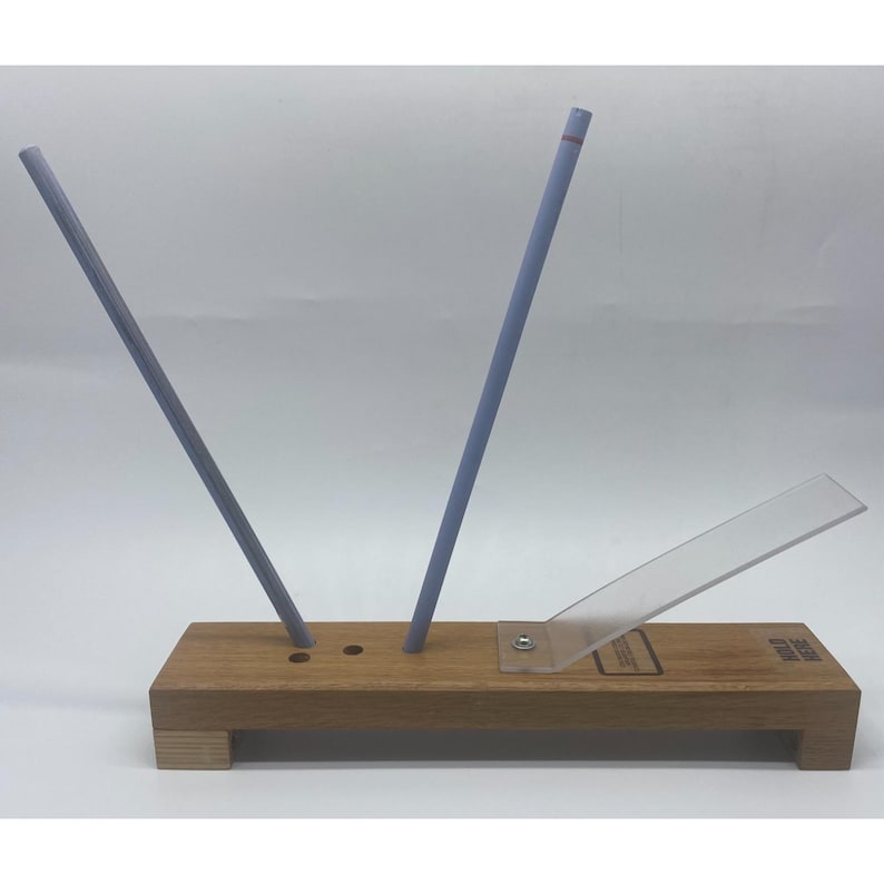 May include: A wooden knife sharpening guide with two angled, light blue sharpening rods. A clear plastic guide is attached to the wooden base. The text "HOLD HERE" is visible on the base.