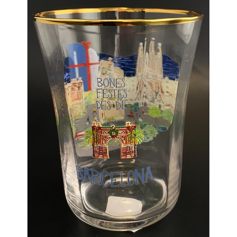 K&ouml;nnte beinhalten: Klares Glas mit Goldrand, verziert mit einer farbenfrohen Illustration von Wahrzeichen Barcelonas. Das Design enth&auml;lt den Text "BONES FESTES DES DE BARCELONA" in Blau und Wei&szlig;, mit Bildern von Geb&auml;uden und einem Torbogen.