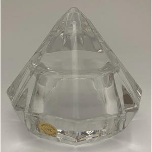 May include: A clear, faceted crystal trinket box with a diamond-like shape. The lid and base are both multi-sided, creating a geometric design. A small, round sticker is affixed to the base.
