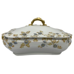 May include: White porcelain serving dish with a lid, adorned with blue and yellow floral patterns. The dish has a gold handle and trim. A decorative piece for serving food.