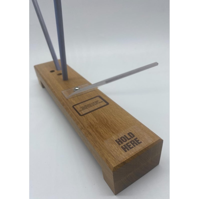 May include: A wooden knife sharpener with three grey sharpening rods and a clear plastic guide. The base is rectangular with the words "HOLD HERE" printed on the side. A small label is also visible.