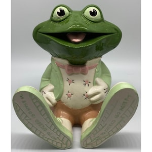 May include: A ceramic frog figurine with a green head and body, wearing a light green jacket, pink bow tie, and tan pants. The frog's shoes are light green with text on the soles. The frog is sitting with its legs extended.