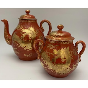 May include: A red ceramic teapot and a matching lidded sugar bowl, both featuring intricate gold dragon illustrations and swirling cloud patterns. Each piece has a rounded body, a curved handle, and a lid with a small knob handle.